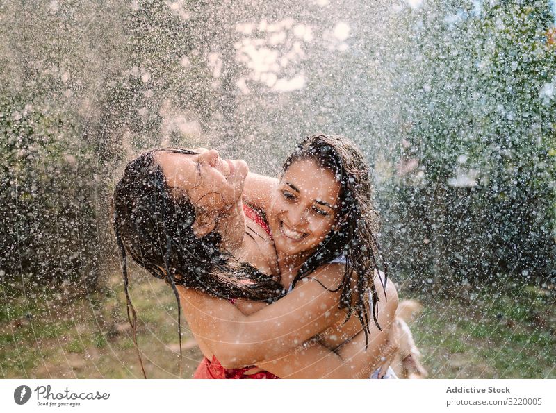 Freunde in Badeanzügen umarmen sich unter Wassertropfen Frauen platschen Garten Tropfen Freundin Umarmung Feiertag fallen Sommer Badeanzug Schlauch Spaß heiter