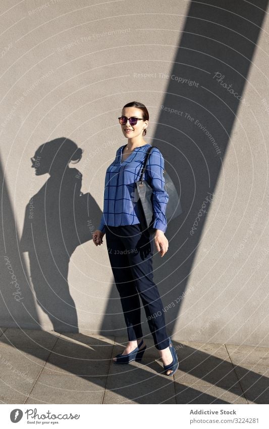 Lächelnde Geschäftsfrau steht an grauer Wand Straße Großstadt Gebäude Straßenbelag elegant modern urban Frau Manager Unternehmer Karriere Tasche Sonnenbrille