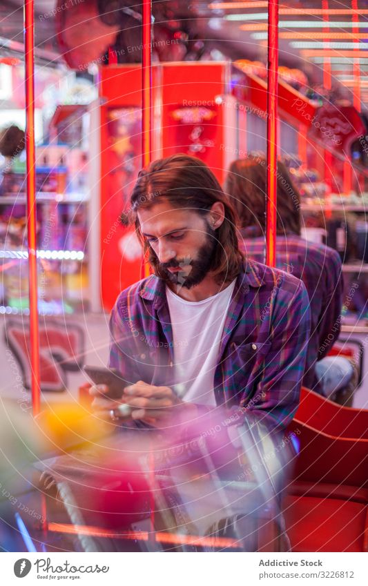 Fokussierter Hipster-Mann simst auf Smartphone, während er sich auf dem Rummelplatz ausruht Texten Messegelände ruhen Karneval fokussiert achtsam bärtig