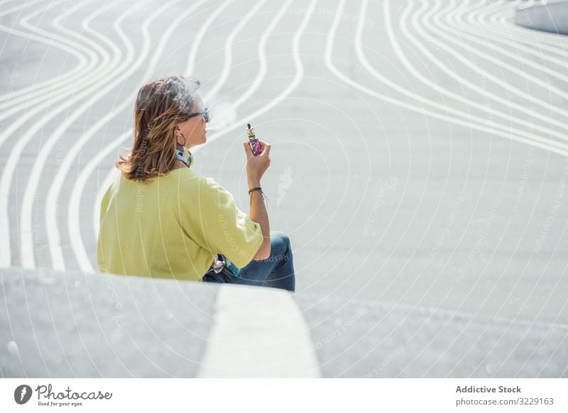 Moderne entspannte Frau, die auf markierter Straße sitzt vaping e Zigarette Rauch Verdunstung räkeln urban Sitzen Kälte Sonnenbrille farbenfroh Dampf Habitus