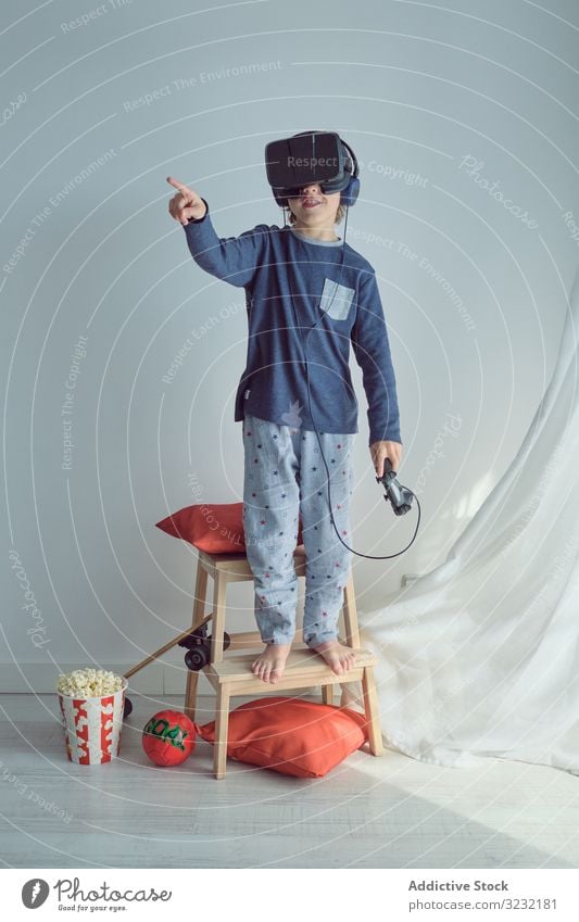 Glückliches Kind im VR-Headset spielen Schutzbrille gewinnen Joystick Tor Sport Popkorn Freizeit Spaß Arme hochgezogen so tun, als ob Spiel Wochenende Simulator