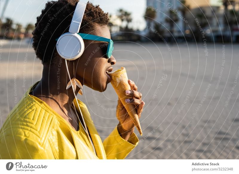 Kühle Frau isst Eiscreme am Strand Speiseeis trendy cool essen wüst Afroamerikaner Kopfhörer Jacke gelb hell Sonnenbrille genießen süß Lebensmittel Sommer Spaß