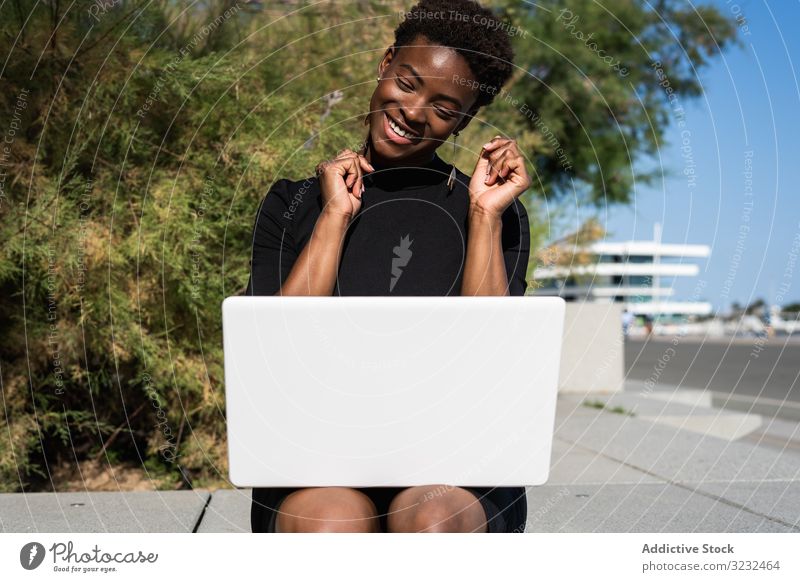 Seriöse Frau surft am Laptop in der Nähe eines modernen Gebäudes sich[Akk] entspannen konzentriert benutzend Surfen Afroamerikaner elegant Straße Kleid schwarz