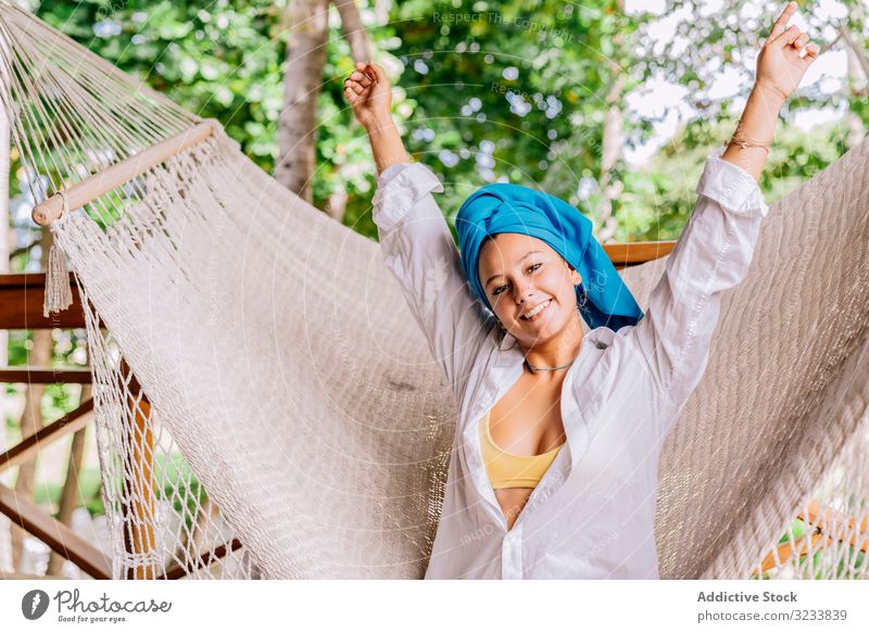 Vergnügte Frau im Turban ruht sich in Hängematte auf Terrasse aus ruhen Glück Lächeln Handerheben sitzen Baum Grün Hemd friedlich Costa Rica jung