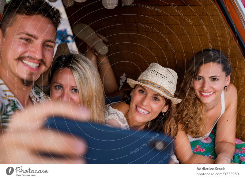 Lächelnder Mann nimmt Selfie mit Freundinnen, die sich bequem auf den Kofferraum eines blauen Autos legen Menschen Selfie nehmen Smartphone PKW Seeküste Freunde