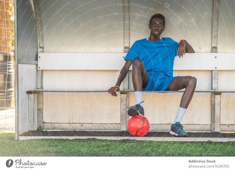 Schwarzer Fussballspieler ruht auf der Bank Teenager Fußball Feld sitzen Pause Ball Training Sportbekleidung ethnisch männlich Jugendlicher sonnig tagsüber