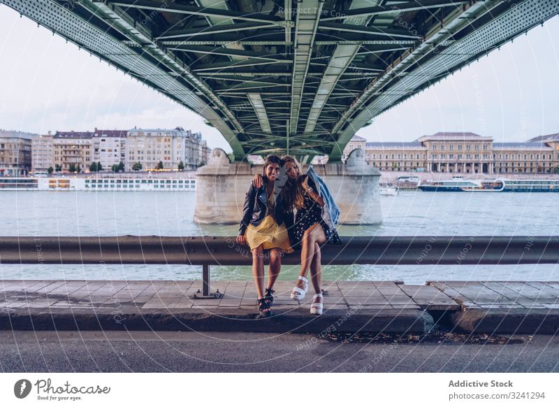 Lächelnde Freundinnen, die sich umarmen und auf dem Zaun entlang des Flusses unter der Brücke sitzen Frauen reisen Großstadt Tourist Architektur Spaß Urlaub