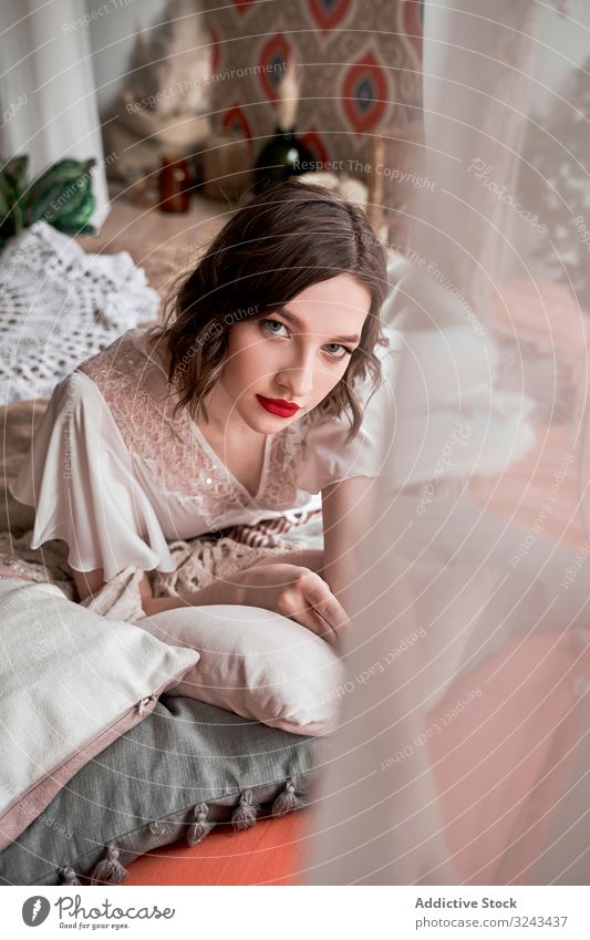 Wunderschöne Frau mit roten Lippen in weißem Kleid, die in die Kamera schaut, während sie auf dem Boden neben dem Sofa sitzt Brautkleid rote Lippen altehrwürdig