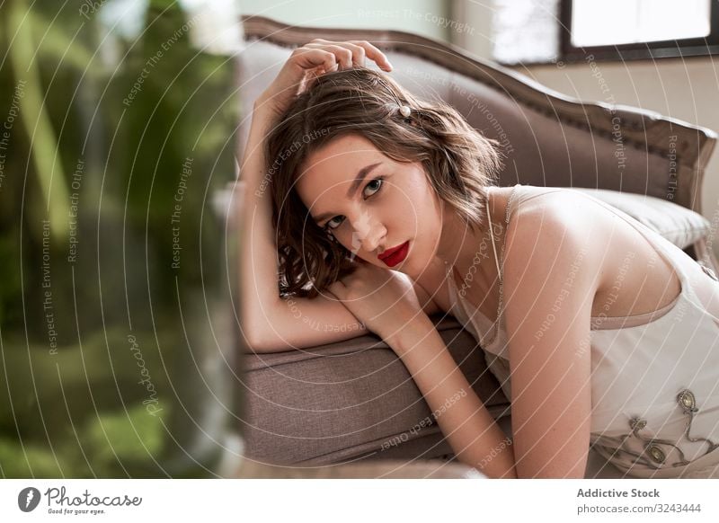 Wunderschöne Frau mit roten Lippen in weißem Kleid, die in die Kamera schaut, während sie auf dem Boden neben dem Sofa sitzt Brautkleid rote Lippen altehrwürdig