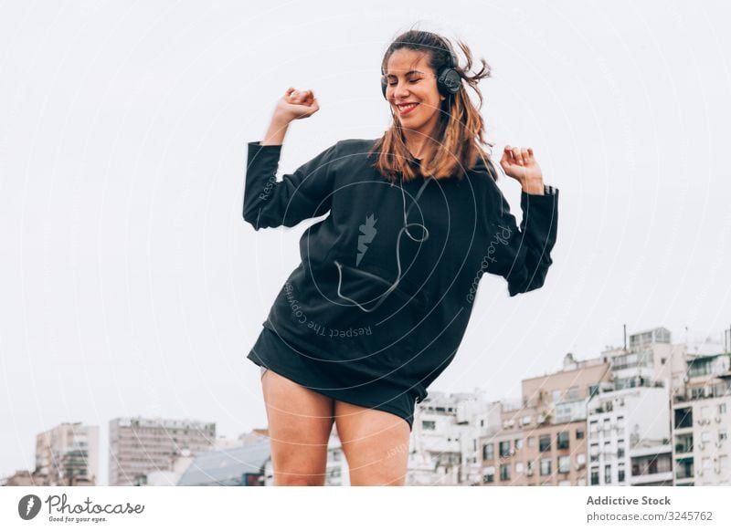 Zufriedene junge Frau in Freizeitkleidung tanzt mit Kopfhörern auf der Straße hören Musik Gerät Tanzen Erhöhung Hände Waffen Gesang Radio Rhythmus Lächeln