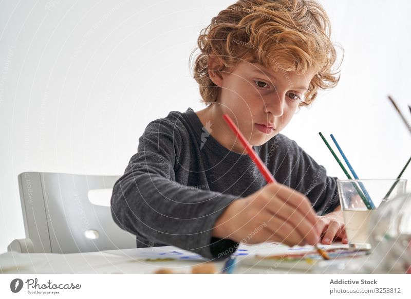 Fröhlicher Junge malt zu Hause mit Aquarellfarben Lächeln Farbe Pinselblume heimwärts Kunst kreativ Bild Bildung Tisch sitzen Kind Werkzeug Handwerk lernen