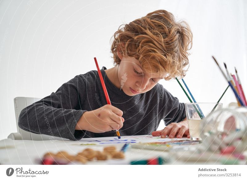 Fröhlicher Junge malt zu Hause mit Aquarellfarben Lächeln Farbe Pinselblume heimwärts Kunst kreativ Bild Bildung Tisch sitzen Kind Werkzeug Handwerk lernen