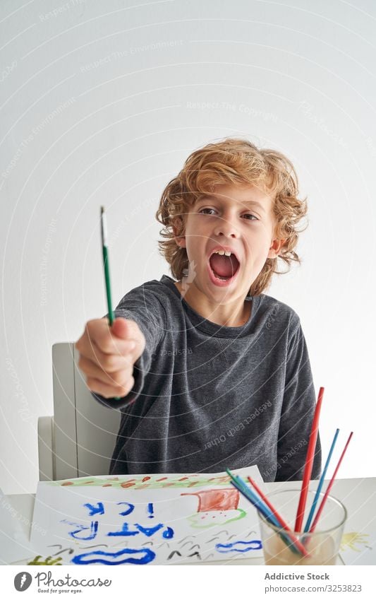 Aufgeregter Junge malt zu Hause Pinselblume aufgeregt schreien zeigen heimwärts Kunst kreativ Bild Wasserfarbe Bildung Tisch sitzen Kind Farbe Werkzeug Handwerk