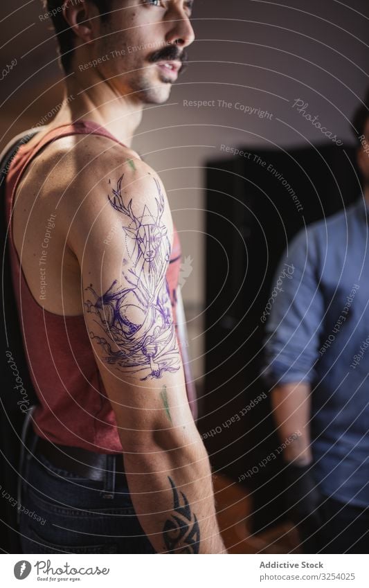 Mann betrachtet Tätowierungsskizze auf dem Arm im Salon Tattoo Arme Aussehen Vorlage Zeichnung prüfen Skizze nachdenken Kunst vorsichtig ernst nachdenklich