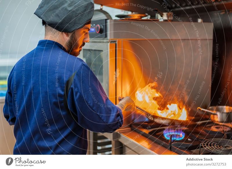 Koch, der mit Flamme in der Pfanne kocht Küchenchef Arbeit Feuer Bratpfanne braten Speise Mann professionell flambieren heiß Herd Gas vorbereiten Uniform Hut