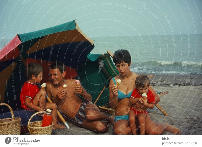 Junge, glückliche Familie sitzt in Badekleidung am Strand, unterm Sonnenschirm und macht Picknick mit Eis, in Italien am Meer, früher in den 60 er Jahren. Sie halten Waffeleis in der Hand und genießen, essen es vor dem Meer.