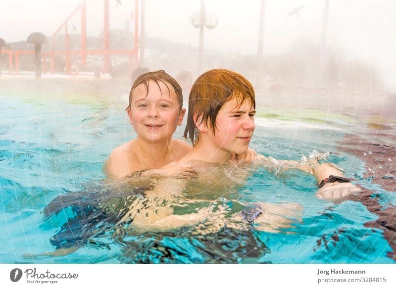 Junge Brüder schwimmen im Außenbereich eines Thermalbads im Winter im warmen Wasser, es ist neblig Freude Glück Spa Schwimmbad Freizeit & Hobby Spielen Sport