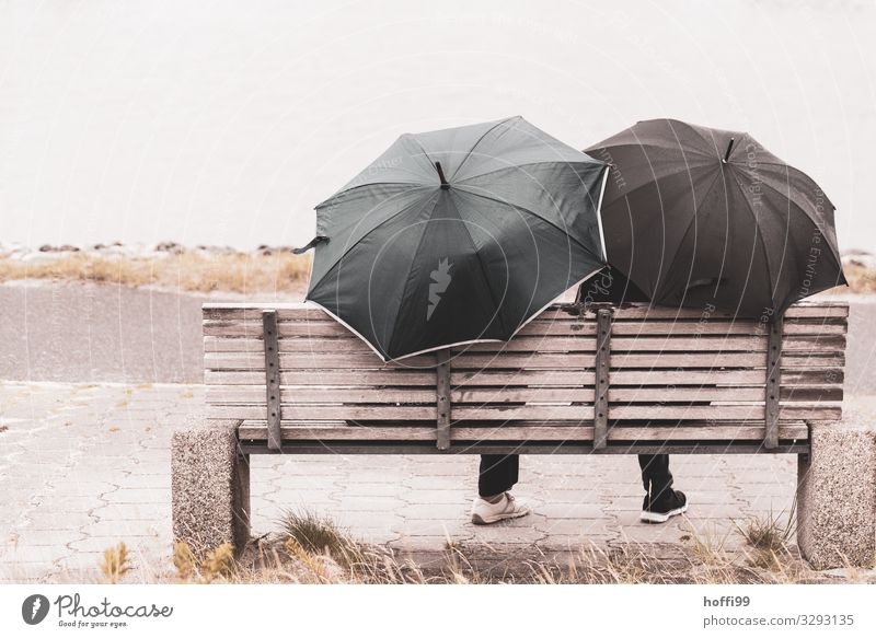 zwei Personen im Regen mit zwei Regenschirmen und jeweils einem sichtbaren Bein auf einer Bank von hinten fotografiert. Erholung ruhig Freizeit & Hobby Ausflug