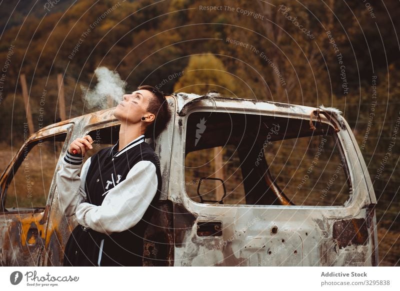 Junge Frau raucht in der Nähe eines beschädigten Autos Zigarette Rauch ausatmen auflehnen PKW rostig Landschaft verbrannt jung fettarm Freiheit Fahrzeug