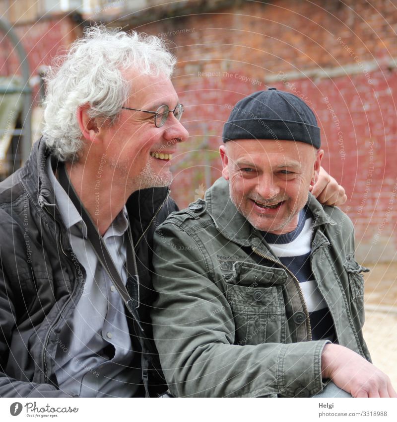 Porträt von zwei lachenden Senioren, einer mit sibergrauen Locken und Brille, der andere mit Mütze vor einer alten Wand Mensch maskulin Mann Erwachsene 2