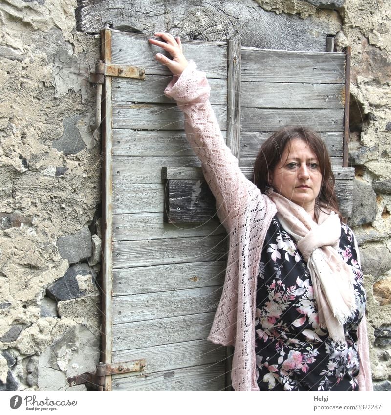 langhaarige brünette Frau mit gemustertem Kleid, rosa Jacke und rosa Schal steht vor einer Wand mit altem geschlossenen Holzfenster Mensch feminin Erwachsene 1