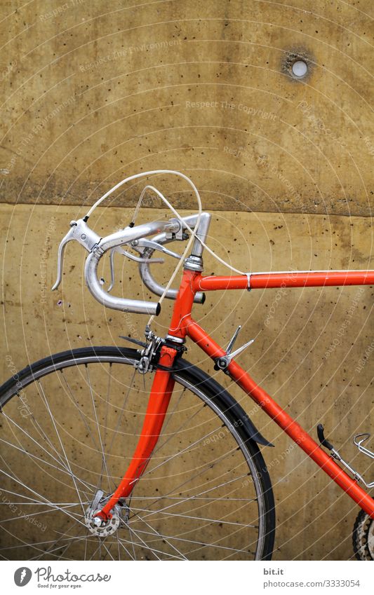 Orangenes Rennrad von einem sportlichen Hipster, steht während einer Pause, geparkt vor einer braunen Wand, im Ausschnitt sind Lenker, Reifen, Vorderrad, Bremse und Gestell des Rades im alten, Retrostil zu sehen.
