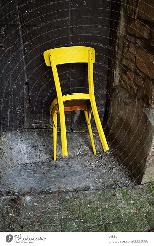 Knallig, gelber, alter, unbenutzter Holzstuhl, steht angelehnt, gekippt, an einer alten Tür aus Holz in einem alten, verlassenen Eingang von einem Haus / Scheune aus Stein.