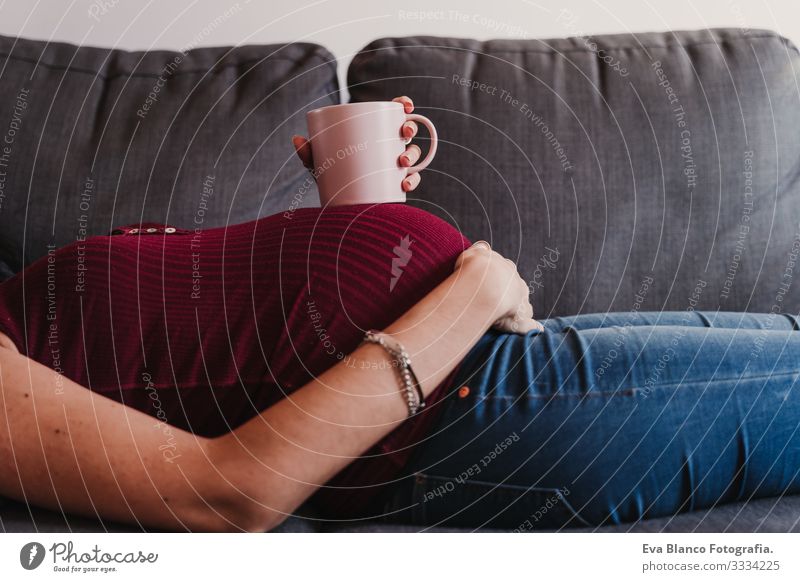 Porträt einer jungen schwangeren Frau zu Hause, die auf dem Sofa liegt und eine Tasse auf dem Bauch hält heimwärts Lifestyle erwartend Fenster Tag Leben
