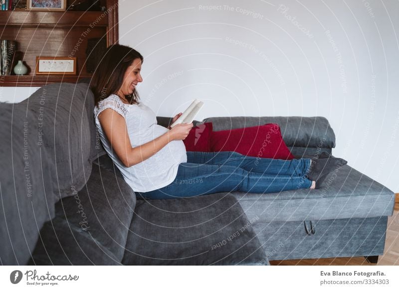 junge schwangere Frau zu Hause, die ein Buch liest Jugendliche Schwangerschaft lesen heimwärts Mutterschaft Leben Lounge bauchfrei Erholung Mitte heiter