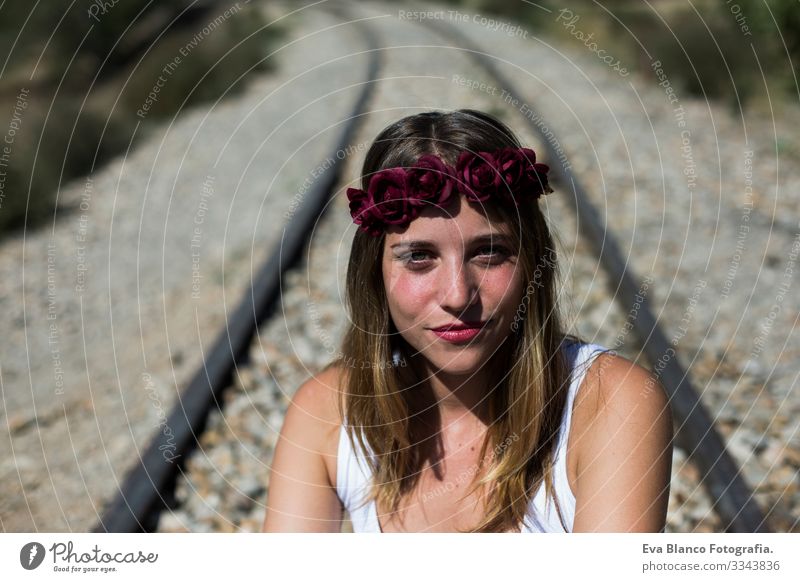 Porträt einer jungen, schönen Frau, die einen roten Rosenkranz auf dem Kopf trägt und in die Kamera schaut, sitzend auf einer Eisenbahn. Im Freien. Sonnig. Lebensstil