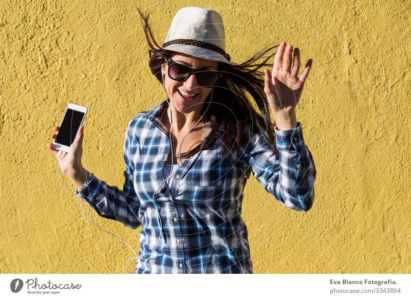 eine glückliche, schöne junge Frau, die Musik hört und sich über dem gelben Hintergrund amüsiert. Sie trägt einen Hut und eine moderne Sonnenbrille. Porträt