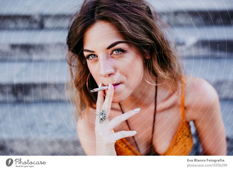 Hübsche junge kaukasische Frau, die an einem sonnigen Tag auf einer Treppe an der Straße der Stadt sitzt und eine Zigarette raucht. Urbaner Lebensstil und Raucherkonzept. Nahaufnahme