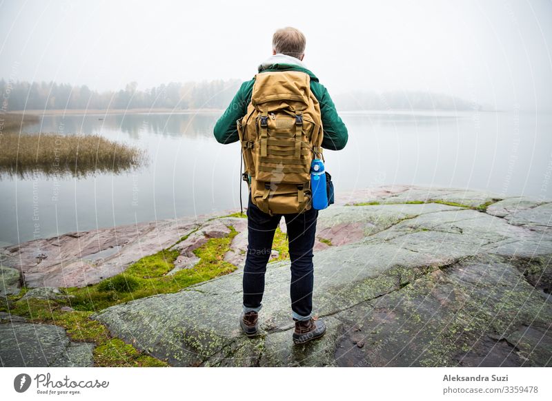 Ein reifer Mann erkundet Finnland im Herbst und schaut in den Nebel. Wanderer mit großem Rucksack auf moosigem Fels stehend. Skandinavische Landschaft mit nebligem Meer und Herbstwald. Rückenansicht