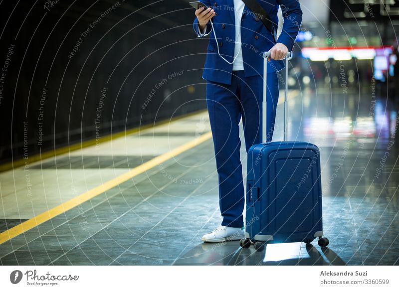Junger, stilvoller, gut aussehender Mann im Anzug mit Koffer steht auf der Metrostation, hält ein Smartphone in der Hand, scrollt und simst, lächelt und lacht.  Vorbeifahrender Zug