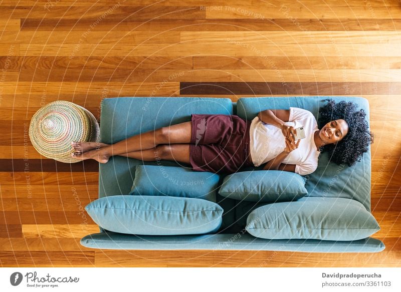 Schwarze Frau chattet auf Smartphone liegend auf Couch zu Hause benutzend Lügen Kälte räkeln Liege Sofa Wohnzimmer modern sich[Akk] entspannen Afroamerikaner