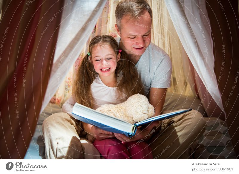 Zeit in Familienqualität. Vater und Tochter sitzen in einem selbstgebauten rosa Zelt mit Blumen, lesen ein großes Buch, sehen sich an, lächeln und lachen. Gemütliches stilvolles Zimmer. Konzept für Familienbande