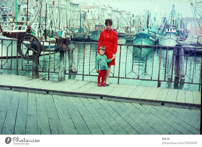 Glückliche, modische, stylische Mutter mit Tochter an der Hand, im Partnerlook, stehen im Urlaub in den 60 er Jahren, während einem Stadtbummel; Ausflug auf einer Brücke im Hafen von Venedig, mit Wasser, Schiffen und Booten im Hintergrund.