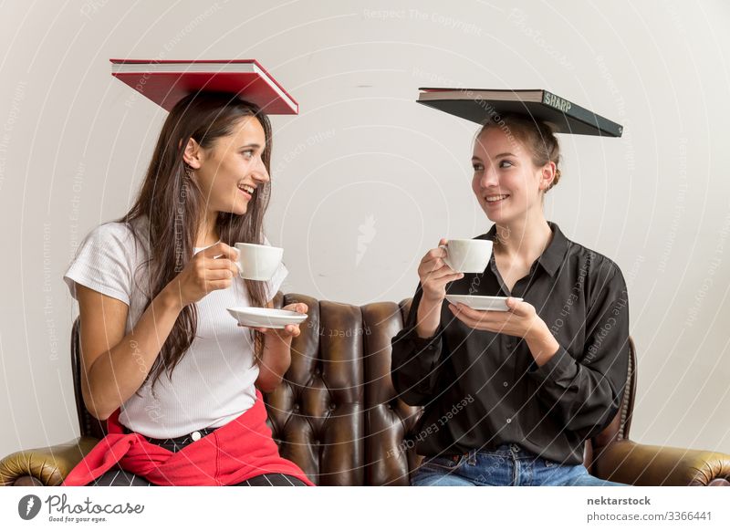 Zwei junge Frauen balancieren Bücher über den Kopf und halten Kaffeetassen, die Augenkontakt herstellen die sich gegenseitig ansehen Mädchen junger Erwachsener