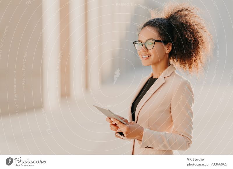 Profilaufnahme einer fröhlichen dunkelhäutigen Frau mit Afro-Haar, die elegant gekleidet ist und einen Tablet-Computer in den Händen hält, auf der Straße spazieren geht und eine optische Brille trägt, die mit Hochgeschwindigkeits-Internet verbunden ist. Lebensstil, Technologie