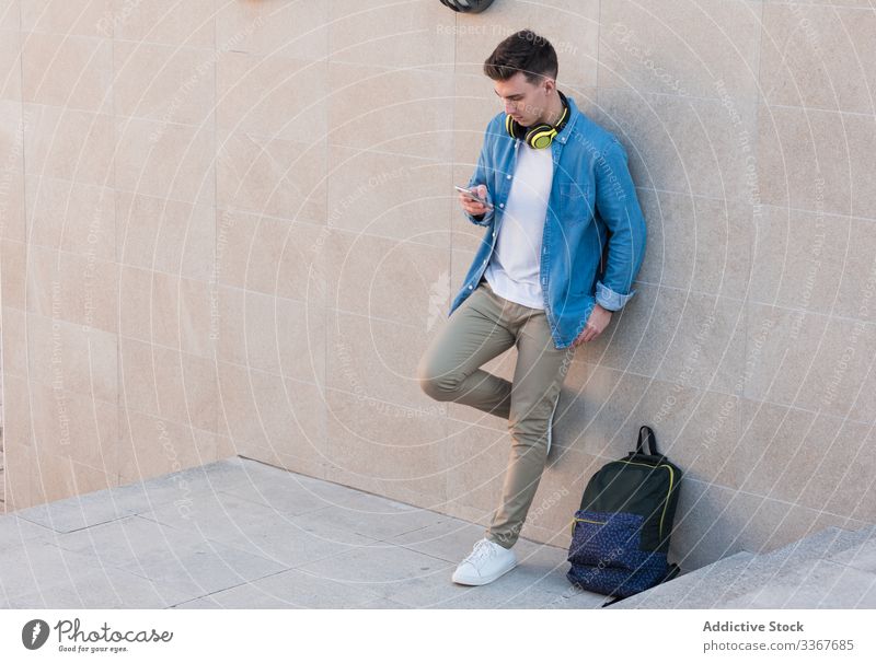 Schüler mit Kopfhörern surft mit einem Smartphone an der Wand lehnend Browsen Rucksack benutzend Bildung klug Mann Apparatur Campus Gebäude Hochschule Surfen