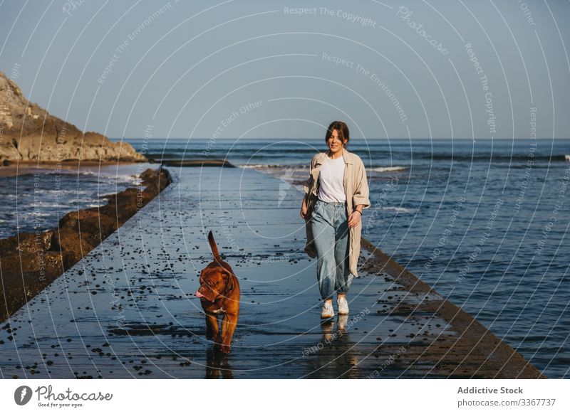 Frau mit Hund beim Spaziergang entlang der nassen Mole gegen ruhiges Meerwasser und felsige Küste bei sonnigem Wetter Pier MEER Wasser Begleiter frisch Haustier