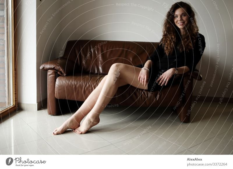 Portrait einer jungen, großen Frau, die barfuß auf einer braunen Leder- Couch sitzt und lächelt Stil Freude schön Leben Wohlgefühl Wohnung Sofa Junge Frau