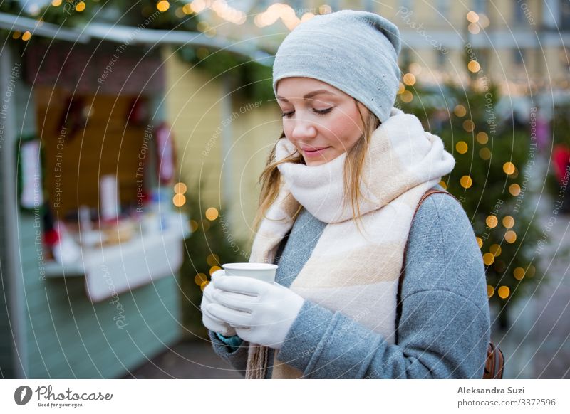 Junge Frau auf dem Weihnachtsmarkt trinkt eine Tasse heiße Schokolade mit Marshmallow und trägt eine warme Strickmütze und einen Schal. Beleuchtete und dekorierte Kioske und Geschäfte im Hintergrund. Helsinki, Finnland