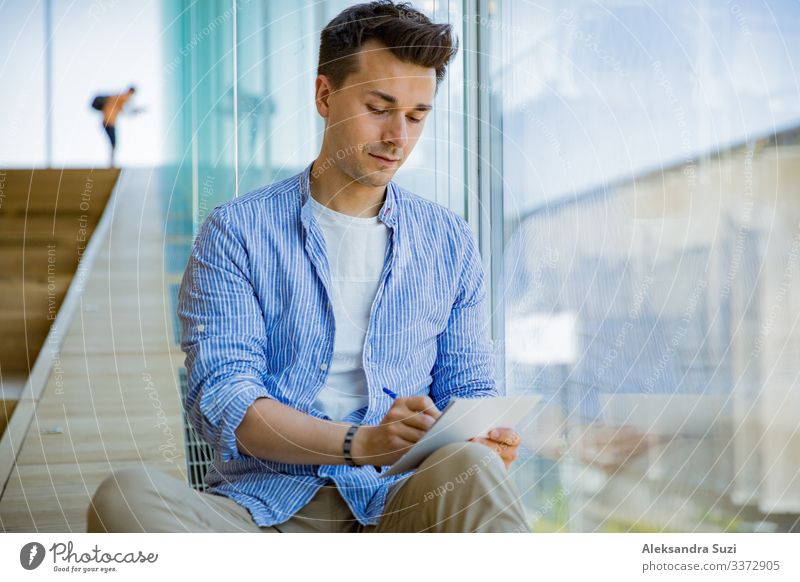 Mann schreibt in einem Notizbuch, sitzt auf Stufen an großen Fenstern, großer Arbeitsraum. Erwachsener Buch lässig Checkliste konzentriert Konzept kreativ