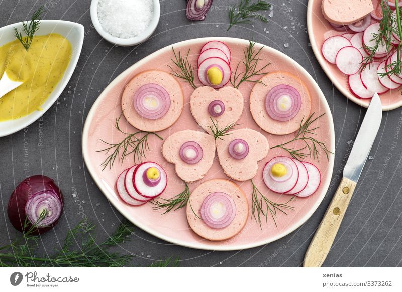 Wurstsalat mit Radieschen, Dill, Zwiebel und Senfsoße auf rosa Teller angerichtet, dazu grobes Salz und Messer mit Holzgriff Wurstwaren Salat Fleisch Essen