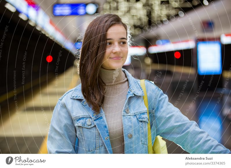 Teenager-Mädchen in Jeans mit Rucksack steht in der U-Bahn-Station, hält Smartphone in der Hand, scrollt und schreibt SMS, lächelt und lacht. Futuristisch helle U-Bahn-Station. Finnland, Espoo