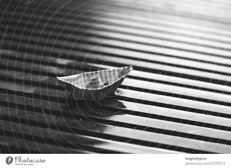 Ein kleines Papierschiff aus Zeitungspapier steht auf einem nassen Holzbrett mit Querrillen Zeitschrift Wasser schlechtes Wetter Regen schwarz weiß Einsamkeit