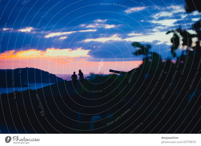 Zwei Frauen unterhalten sich beim Sonnenuntergang auf einer Insel. Dunkler Vordergrund mit Kanone. Blaue Wolken und mit Blick auf eine Nachbar Insel. Die Frauen sind nur als Schatten zu erkennen.