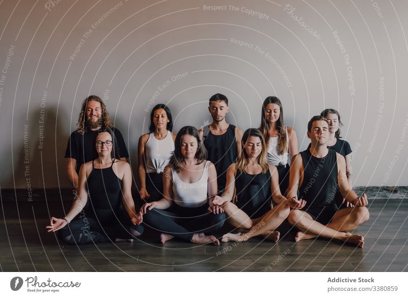 Gruppe von ruhigen, entspannten Athleten, die zusammen Yoga praktizieren und in einem hellen, modernen Trainingsraum eine Lotus-Pose ausführen meditieren