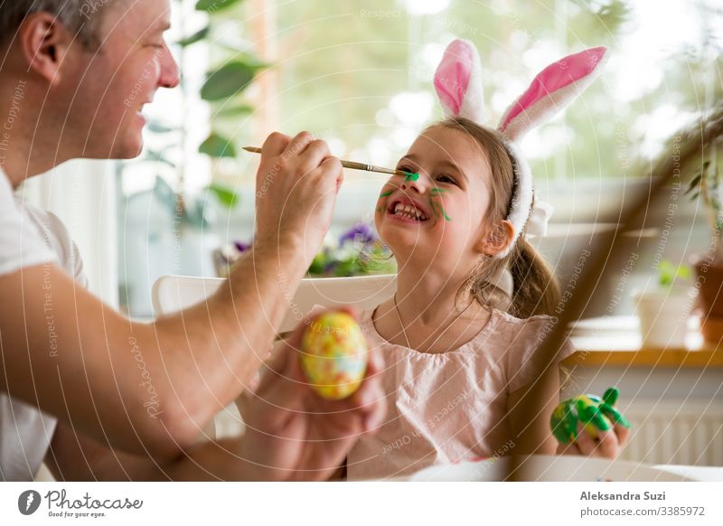 Ein Vater und eine Tochter feiern Ostern und bemalen Eier mit dem Pinsel. Eine glückliche Familie, die lächelt und lacht, die sich auf das Gesicht gemalt hat. Ein süßes kleines Mädchen in Hasenohren, das den Feiertag vorbereitet.